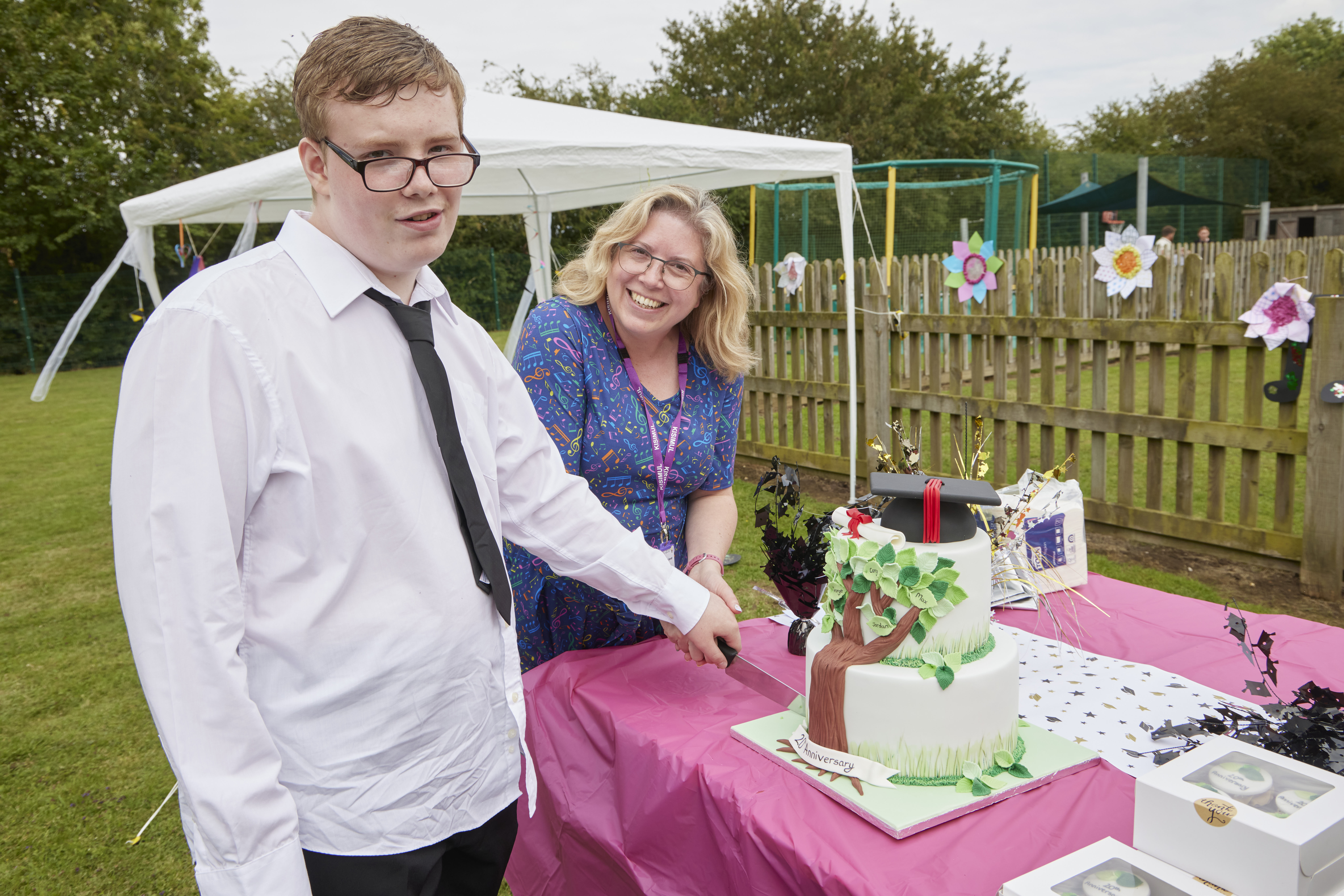 A young man and woman cutting a graduation cake at an outdoor event.