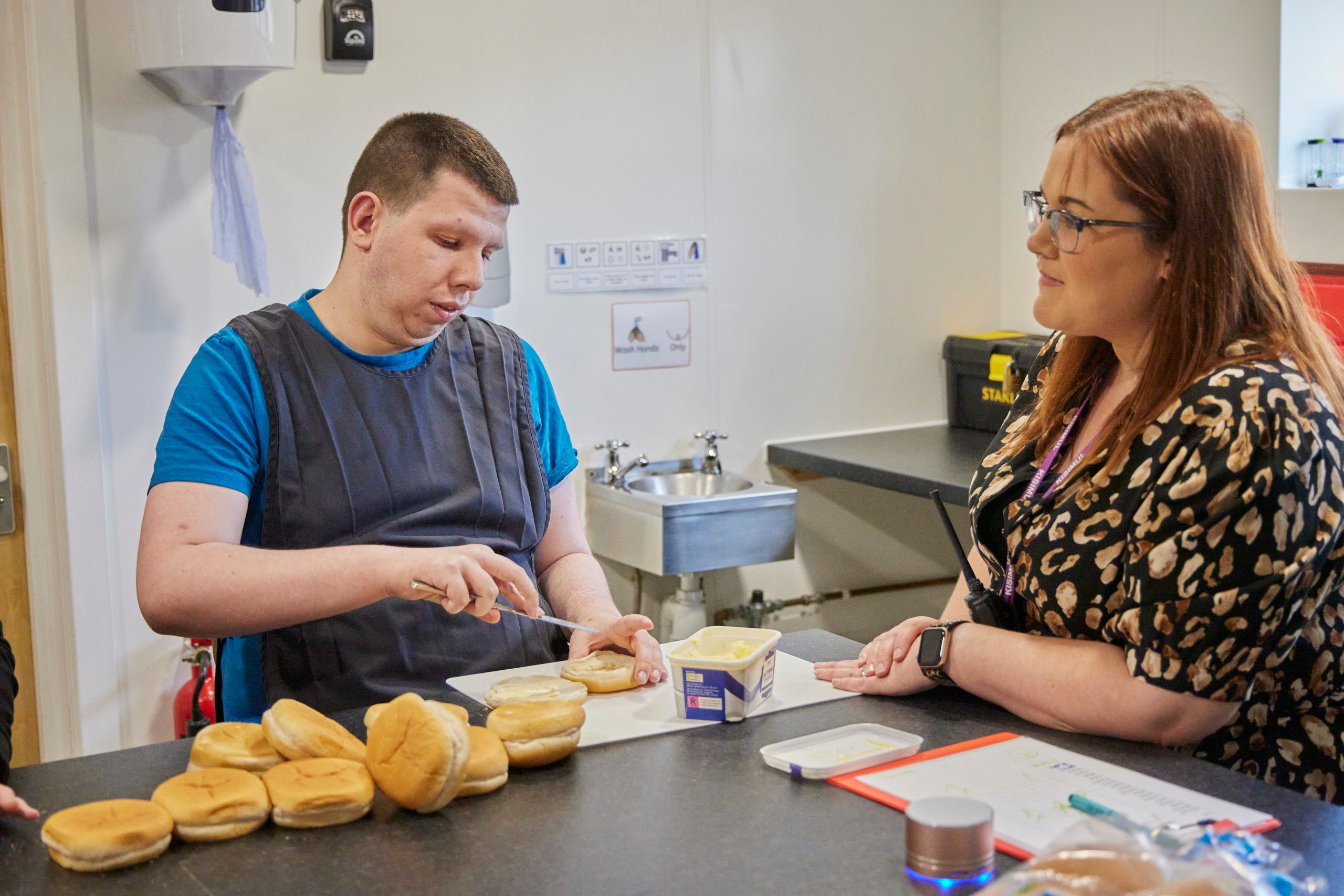 Man spreading butter on buns with a woman watching. Kitchen setting.