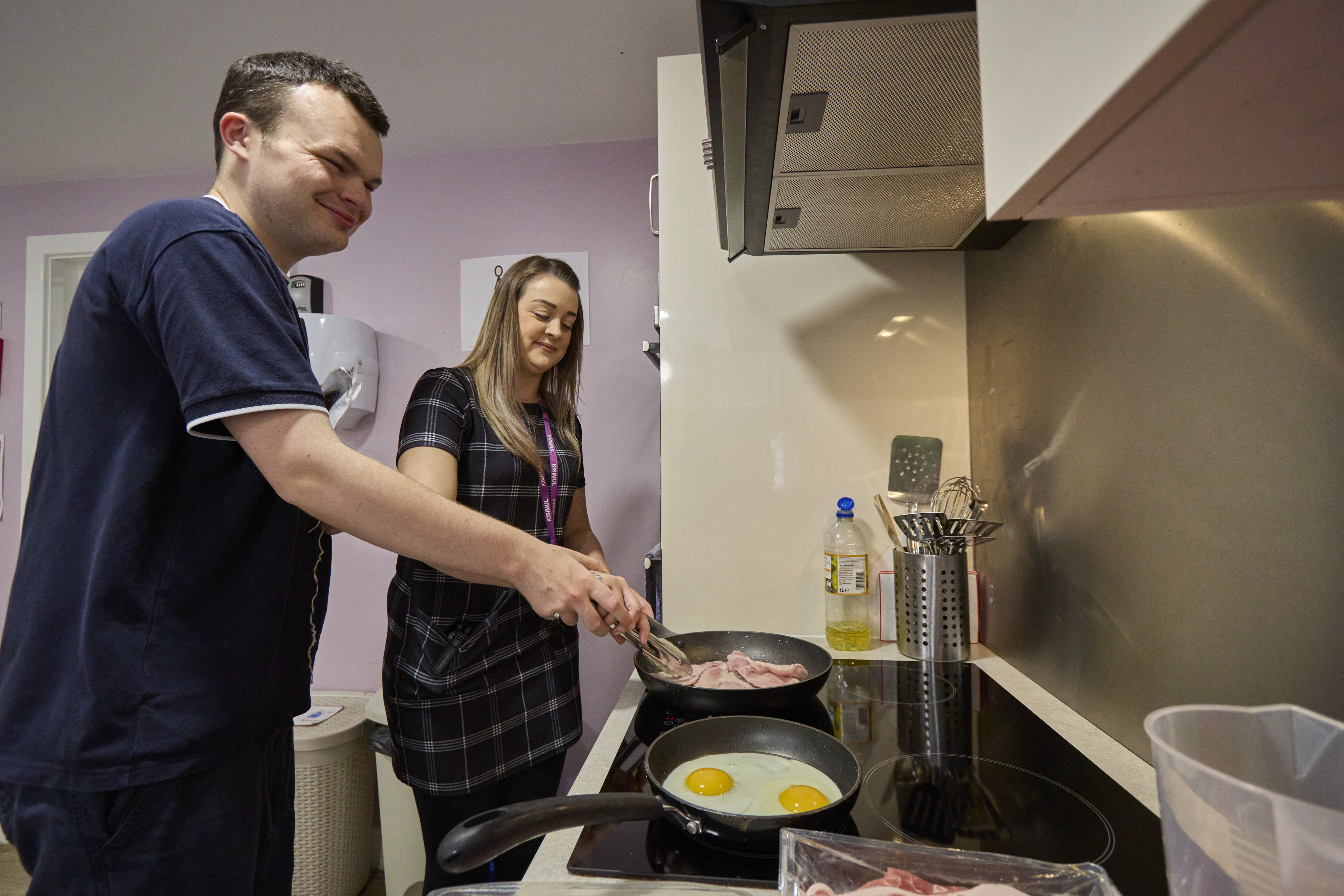 Two people cooking eggs and bacon in a kitchen.