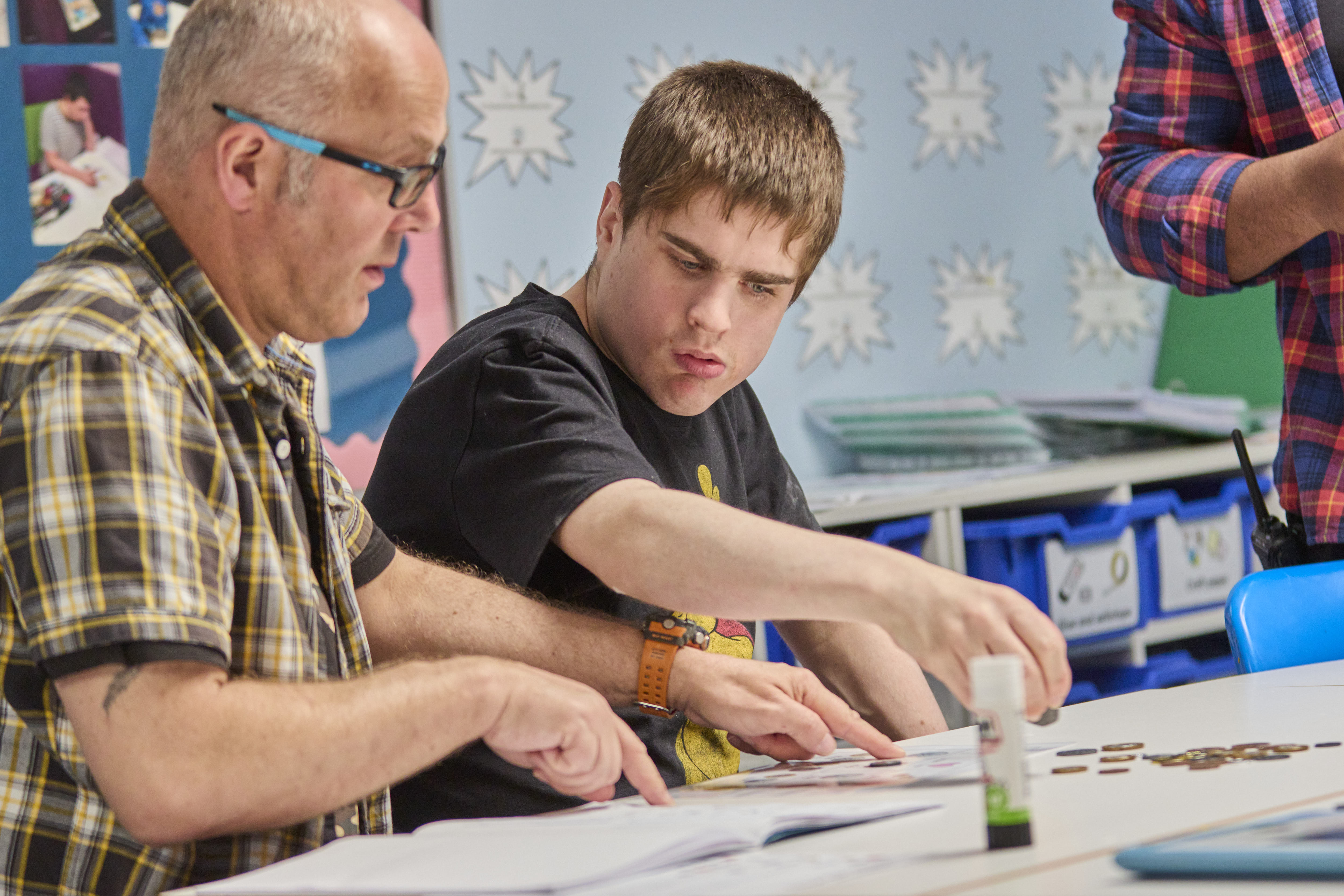 A teacher in a plaid shirt points to a page as a student works on a classroom activity.