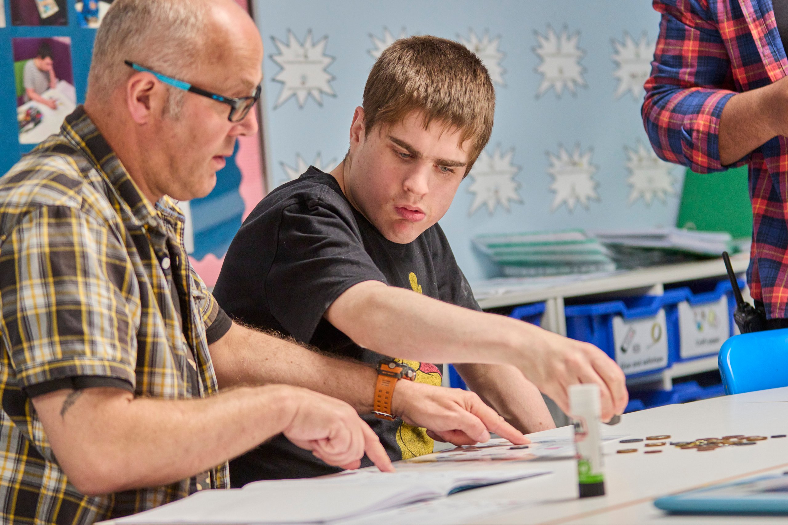 A teacher in a plaid shirt points to a page as a student works on a classroom activity.